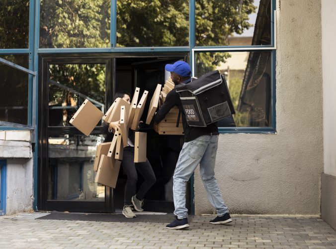 African American courier struggles with a heavy pile of pizza boxes during a takeaway delivery. Uncoordinated poorly trained worker ruining the meal order, bad service with the stack falling.