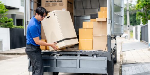 An Asian delivery man or mover in a blue uniform is unloading cardboard boxes from his truck. cargo, courier, freight, shipping service, house moving service