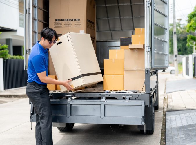 An Asian delivery man or mover in a blue uniform is unloading cardboard boxes from his truck. cargo, courier, freight, shipping service, house moving service