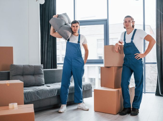 Busy day. Two young movers in blue uniform working indoors in the room.