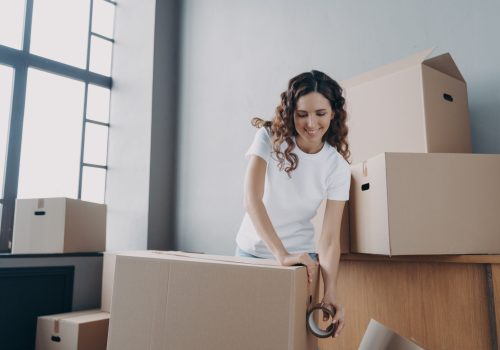 Happy girl moves alone. Young woman is packing boxes with adhesive tape. Lady is wrapping cardboard boxes with packing tape. Moving service worker preparing boxes for shipping and storage.