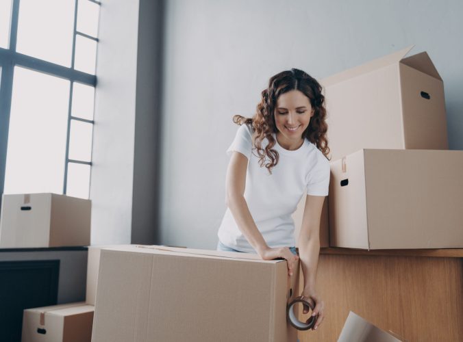 Happy girl moves alone. Young woman is packing boxes with adhesive tape. Lady is wrapping cardboard boxes with packing tape. Moving service worker preparing boxes for shipping and storage.