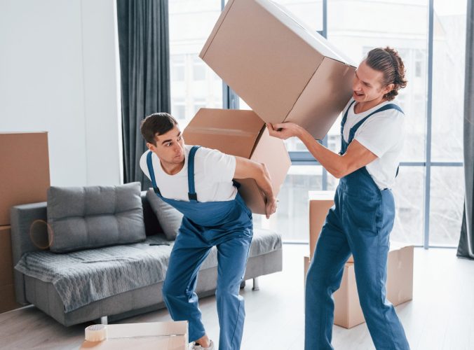 Heavy boxes. Two young movers in blue uniform working indoors in the room.
