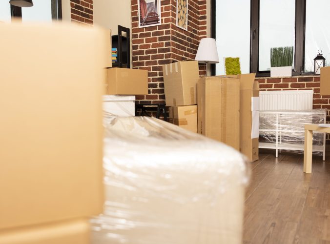 Unfurnished living room with cardboard boxes and scattered furniture near brick wall. Empty apartment prepared for moving day with packages, wrapped sofa and tables awaiting new tenants or owners.